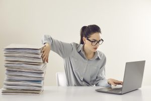 Young accountant working on laptop computer sitting at desk with pile of papers. Paperwork vs electronic documents. Storing files in digital database. Having quick convenient access to storage system